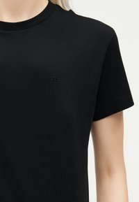 Μαύρο κοντομάνικο μπλουζάκι με στρογγυλή λαιμόκοψη, που φοριέται από άτομο, με μικρό μαύρο κεντημένο «R» στο στήθος.