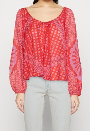 Femme portant un chemisier rouge à manches longues avec des motifs floraux et géométriques roses, associé à un jean bleu clair.