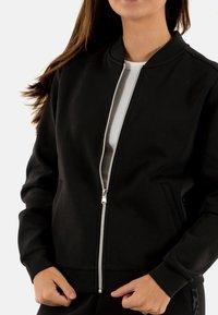 Giacca nera con zip realizzata in tessuto liscio, con colletto a costine, tasche laterali e dettagli con zip metallica. Fodera grigio chiaro visibile.
