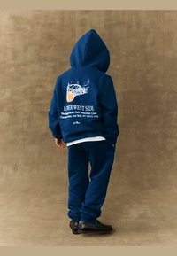 Tumši zilas krāsas kapuce un atbilstoši sporta bikses ar baltām grafikām, kas attēlo basketbola grozu un tekstu uz muguras. Audums šķiet mīksts un ar tekstūru.