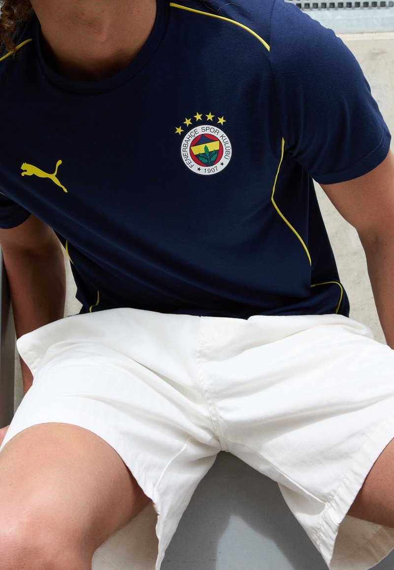 Camiseta deportiva azul marino de Puma con detalles en amarillo y logo de Fenerbahçe; combinada con pantalones cortos blancos. Textura suave, diseño atlético casual.
