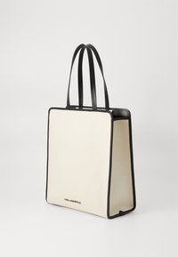 Krämfärgad canvas tote-väska med svart läderdetalj, rektangulär form, två stadiga handtag och en tryckt logotyp på den nedre fronten.