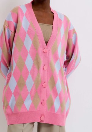 Cardigan rose ample avec motif en losanges bleu clair et beige, cinq boutons roses, porté sur un haut beige et un pantalon beige.
