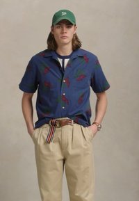 Κοντομάνικο ναυτικό μπλουζάκι με κόκκινες και πράσινες σχέσεις, σε συνδυασμό με μπεζ παντελόνι και πολύχρωμη ριγέ ζώνη. Πράσινο καπέλο στο κεφάλι.