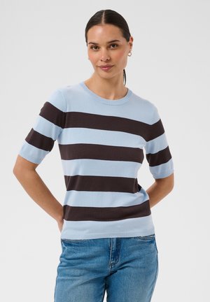 Kvinde med mørkt hår bundet tilbage iført en lyseblå og mørkebrun stribet kortærmet sweater og blå jeans, stående med hænderne bag ryggen.