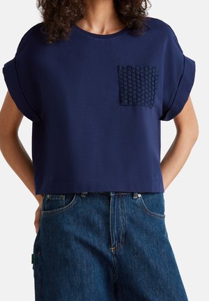 Kvinde iført en navyblå t-shirt med korte ærmer, med en tekstureret brystlomme og løstsiddende blå denimjeans.