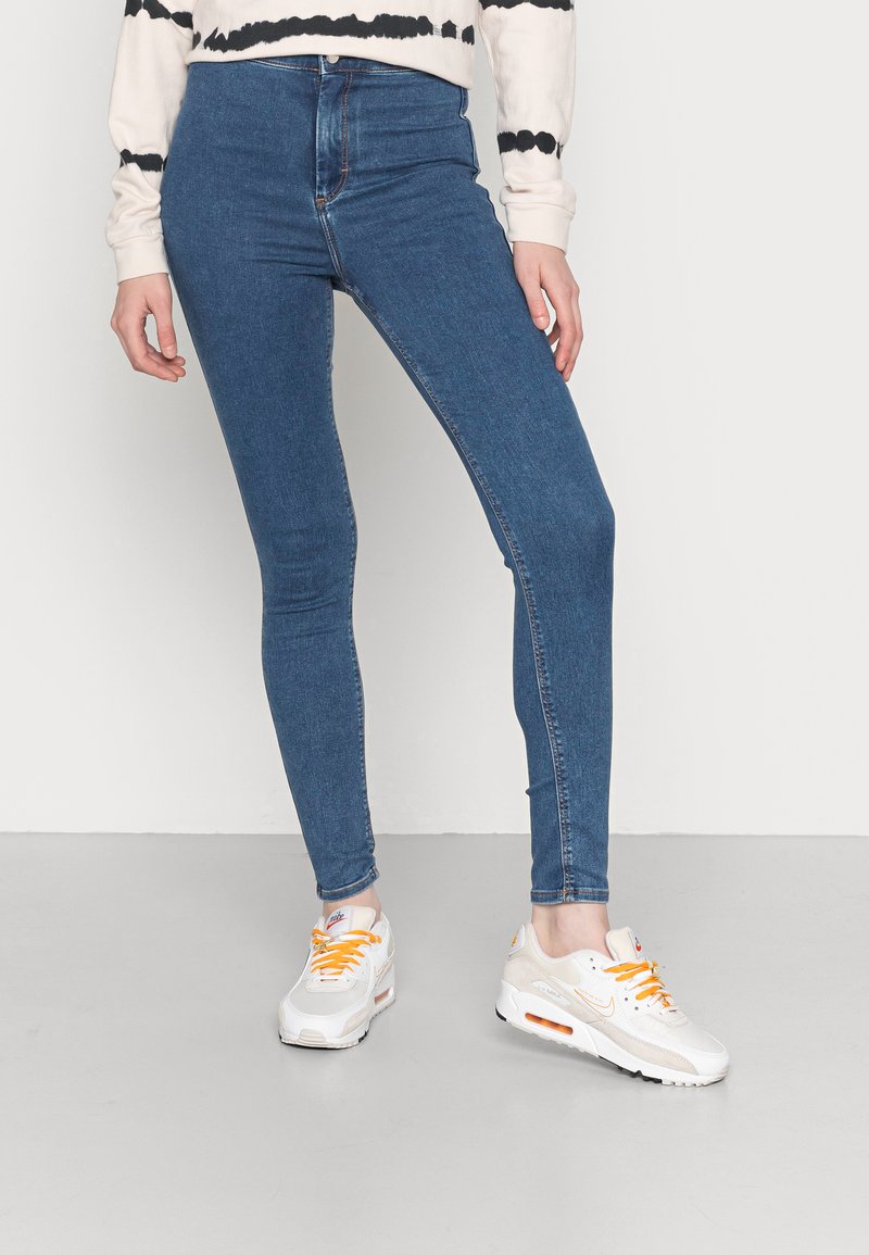 Jeans skinny de denim azul com alta cintura e design de cinco bolsos, acompanhados por ténis brancos com atacadores e detalhes em laranja.