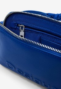 Modrá kožená taška so strieborným zipsom, s textúrovaným povrchom a malou vnútornou kapsou. Názov značky je vytlačený na boku.