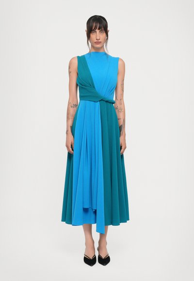 Ärmelloses Kleid in Blau- und Türkistönen, mit einem verdrehten Oberteil, plissiertem Rock und fließender Silhouette. Getragen mit schwarzen High Heels.