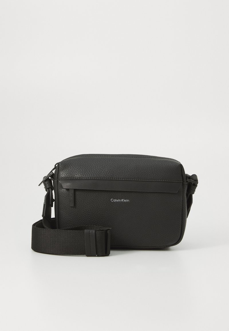 Black leather crossbody bag with textured surface, top zipper, adjustable strap, and embossed Calvin Klein logo on the front.