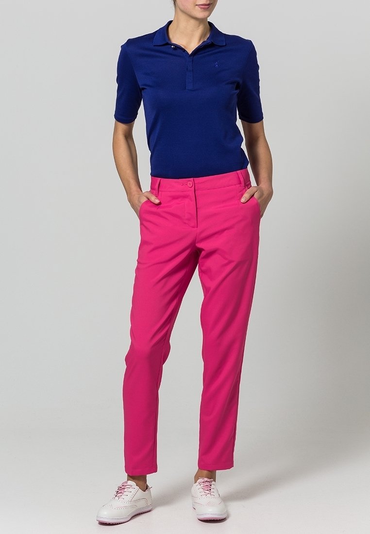 Kobieta stojąca z rękami w kieszeniach, ubrana w granatową koszulkę polo, jaskrawo różowe spodnie oraz białe trampki z różowymi sznurowadłami.