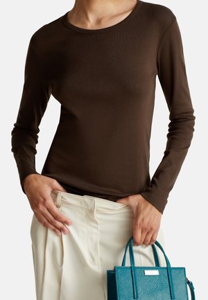 Person trägt ein braunes langärmeliges Oberteil, das in cremefarbene Hosen gesteckt ist, und hält eine strukturiere türkisfarbene Handtasche an den Griffen.
