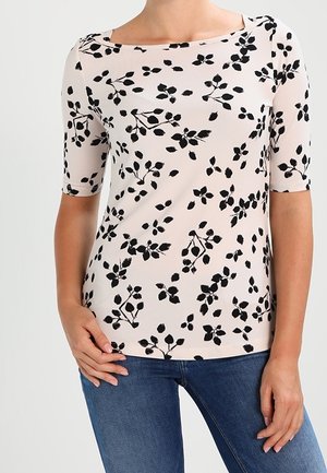 Kvinde iført en lyserød kortærmet top med sort blomsterprint og blå jeans, stående med den ene hånd på hoften og den anden ned langs siden.