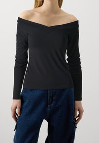 Black off-the-shoulder long-sleeve top, featuring a v-neckline and slim fit. Soft fabric with minimal texture, paired with blue denim pants.
