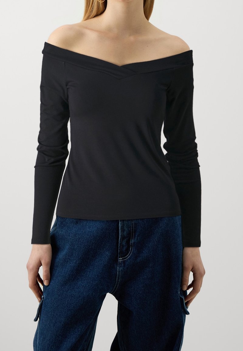 Black off-the-shoulder long-sleeve top, featuring a v-neckline and slim fit. Soft fabric with minimal texture, paired with blue denim pants.