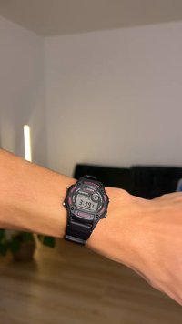 Digitalni ručni sat s kućištem i remenom od crne smole. Ima digitalni zaslon s vremenom i datumom, osvijetljeno dugme označeno s "SVJETLO".