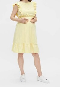 Abito premaman giallo con spalle arricciate, orlo a strati e maniche corte. Abbinato a sneakers bianche. Tessuto semplice.