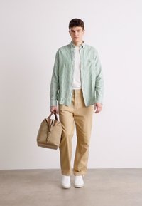 Camicia button-up a righe verdi e bianche, pantaloni beige, sneakers bianche e una borsa beige con zip. Design semplice e moderno con colori distintivi.
