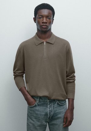 Young man with short curly hair wearing a brown long-sleeve polo shirt with a zipper and light blue jeans, standing against a light wall.