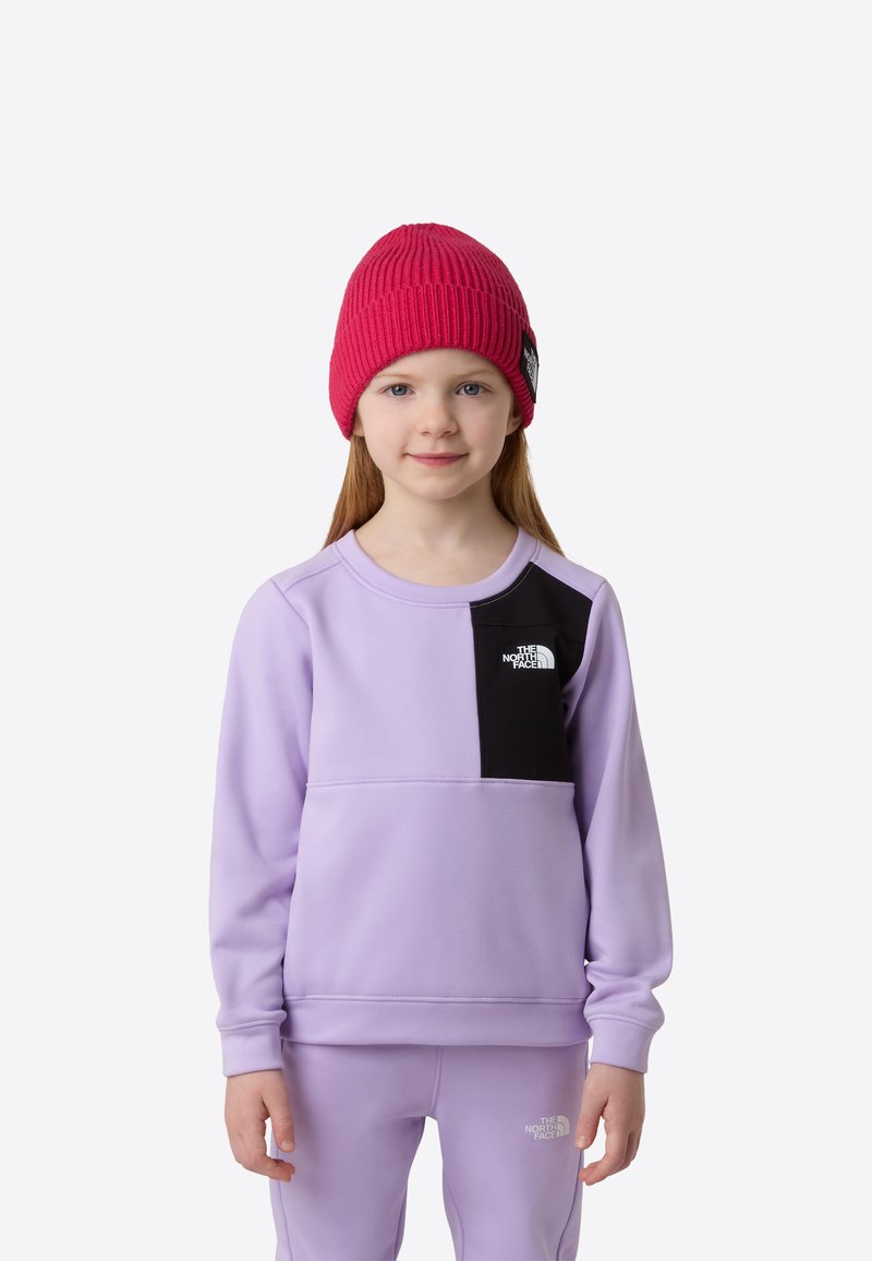 Lila Sweatshirt mit schwarzem Einsatz und Logo, kombiniert mit passenden lila Hosen. Rote, gerippte Mütze sitzt auf glattem, braunem Haar.