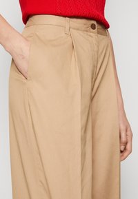Pantalon beige sur mesure avec plis, doté d'une fermeture à bouton et de poches latérales. Le tissu semble doux avec une texture lisse.