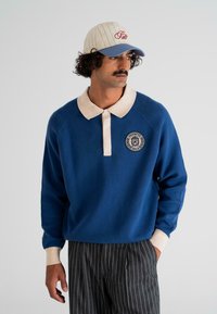Man wearing blue long-sleeve polo with beige collar and cuffs, gray pinstripe trousers, and white baseball cap with blue brim, looking right.