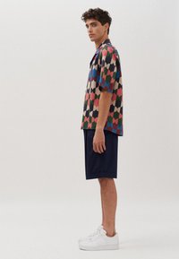 Bunt gestreiftes Kurzarmhemd mit geometrischem Muster in Rot, Blau, Grün und Beige, kombiniert mit marineblauen Shorts und weißen Turnschuhen.