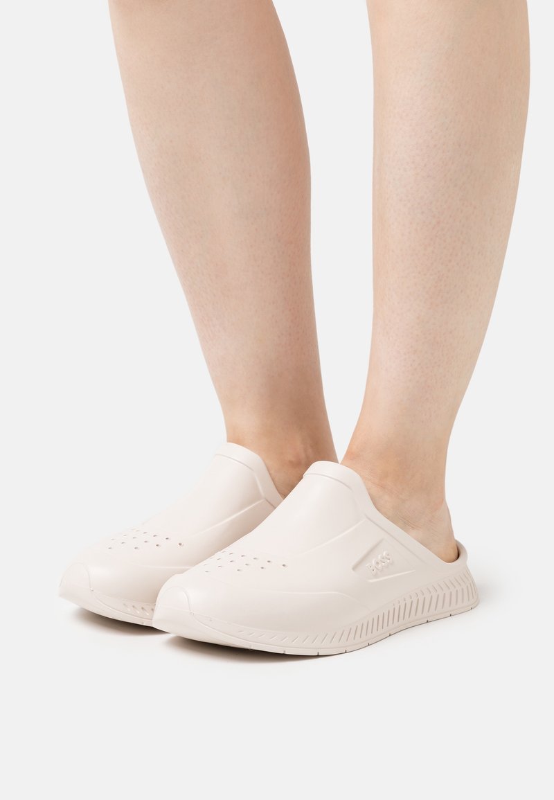 Personne portant des sabots légers beige clair à enfiler avec des trous de ventilation, debout sur une surface blanche.