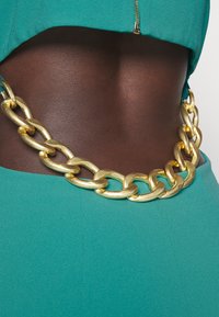 Zlatý řetězový pás s velkými články, s texturovaným povrchem, nošený kolem tyrkysového oděvu. Pás dodává outfitu kovový akcent.