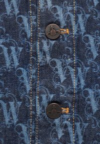 Kék tónusú monogramos minta a farmer anyagon. Két sötét gomb logóval és sárga öltésekkel, amelyek a varráson láthatók.