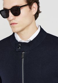 Giacca zip-up navy con colletto alto, tessuto testurizzato, due bottoni neri e una zip argentata. Indossata con occhiali da sole neri e una camicia bianca.