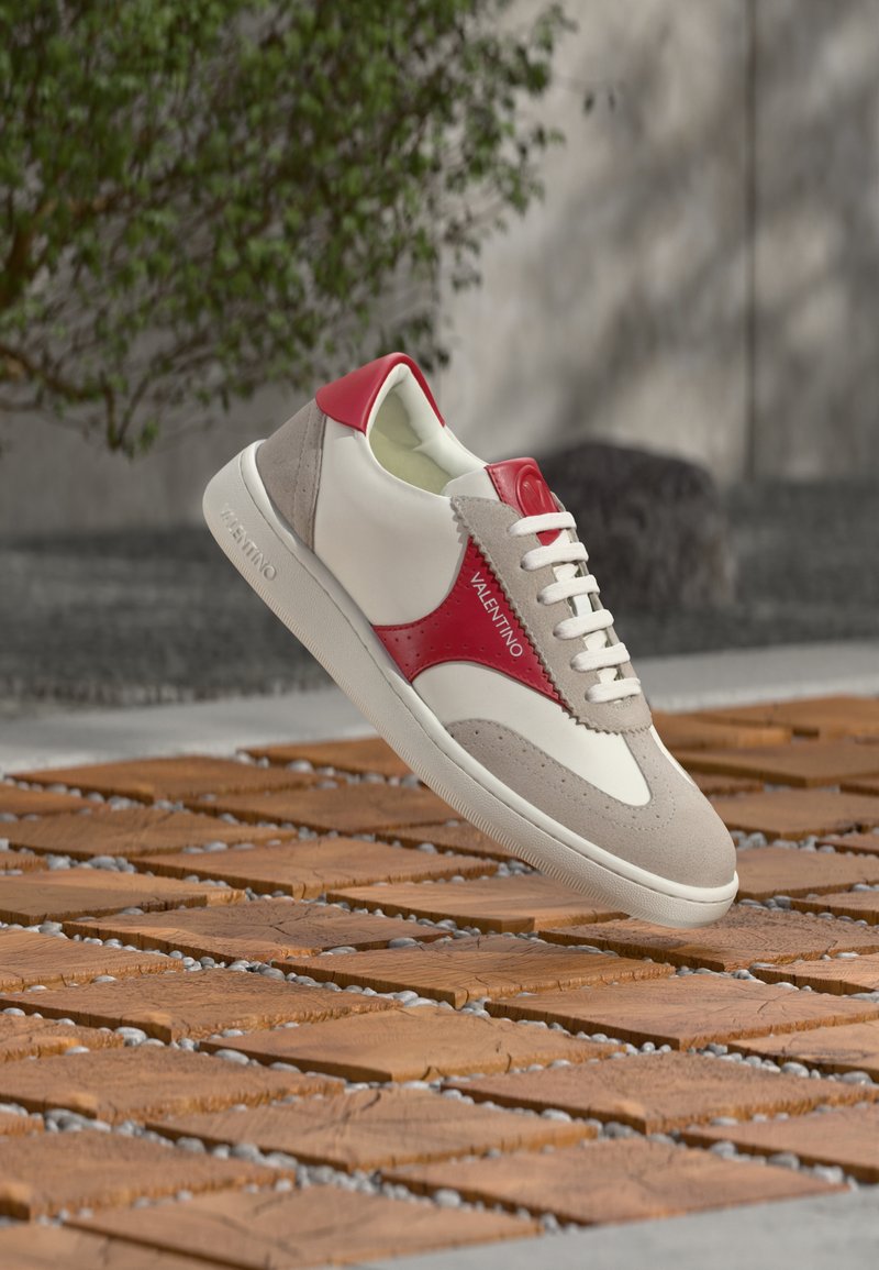 Baskets Valentino blanches et grises avec des accents rouges, suspendues au-dessus de carreaux en bois et de galets gris en plein air près d'un buisson vert.