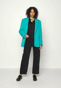 Blazer oversized em turquesa, camisa preta com botões, jeans largos pretos e tamancos pretos. Cabelo cacheado, braços relaxados ao lado.