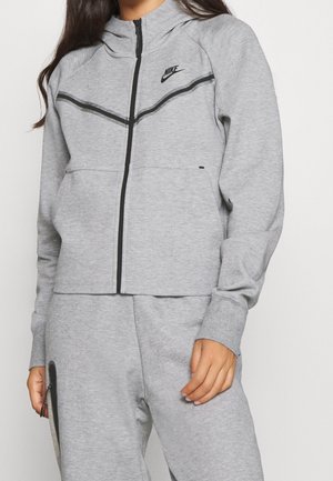 Personne portant un survêtement Nike gris clair avec un sweat à capuche zippé et un pantalon assorti, avec des détails noirs et un logo Nike sur la poitrine.