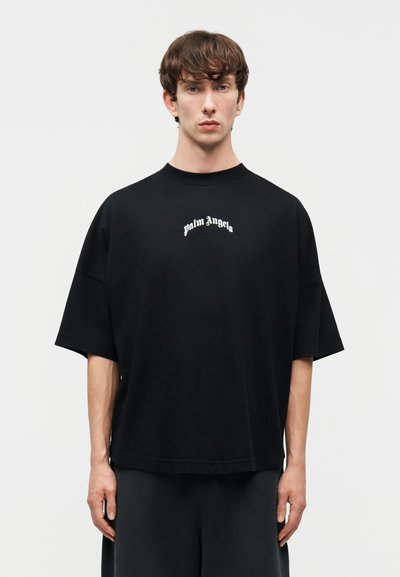 Noor mees lühikeste pruunide juustega kannab musta üle suurusega T-särki, mille rinnal on valge kiri "palm angels", seistes lihtsa helehalli tausta ees.