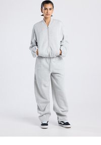 Vaaleanharmaa yläosan ja housujen muodostama ylisuurikokoinen setti pehmeästä kankaasta. Ylhäosan etuosassa on vetoketju ja resorit hihansuissa; housuissa on joustava vyötärö ja taskut.