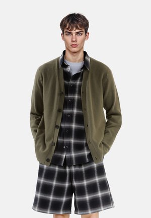 Cardigan olive vert porté sur une chemise à carreaux noir et blanc, associé à un short à carreaux assorti. La texture lisse et les détails des boutons sont visibles.