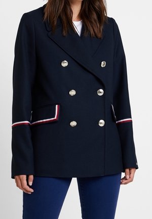 Femme portant un blazer croisé bleu marine avec des boutons argentés et un passepoil rayé rouge, blanc et bleu sur les poches et les manches.