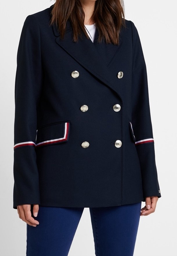 Femme portant un blazer croisé bleu marine avec des boutons argentés et un passepoil rayé rouge, blanc et bleu sur les poches et les manches.