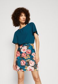 Türkise Bluse mit kurzen Ärmeln, kombiniert mit einem figurbetonten Blumensock, der große rosa Rosen auf einem blauen Hintergrund und kleine gelbe Akzente zeigt.