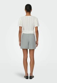 Witte gebreide top met korte pofmouwen, versierd met opengebreide details. Gecombineerd met lichtgrijze shorts en zwarte instapschoenen.