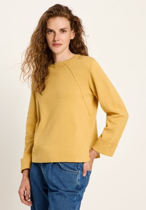 Frau mit langen lockigen Haaren, die einen senfgelben Pullover und blaue Jeans trägt, posiert mit einer Hand in der Tasche vor einem hellen Hintergrund.
