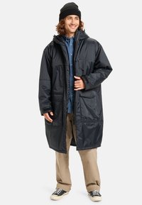 Zwarte waterdichte lange jas met een capuchon, grote voorzakken en ritssluiting. Getextureerde stof en een relaxte pasvorm. Gekocht over een blauwe shirt en khaki broek.