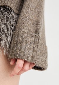 Brun strikket genserarm med ribbestrikket mansjett; myk, teksturert stoff med en blanding av lyse og mørke fibre; hånd som hviler nær kanten.