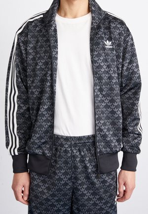Mand iført sort Adidas træningsdragt med gråt trebladsmønster og hvide striber på ærmerne, kombineret med en almindelig hvid t-shirt.
