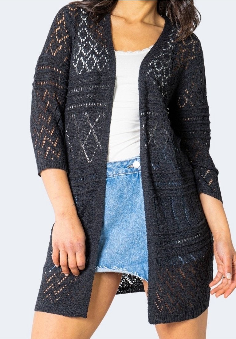 Femme portant un cardigan noir en maille ajourée sur un haut blanc et une jupe en denim bleu clair, debout les mains détendues le long du corps.