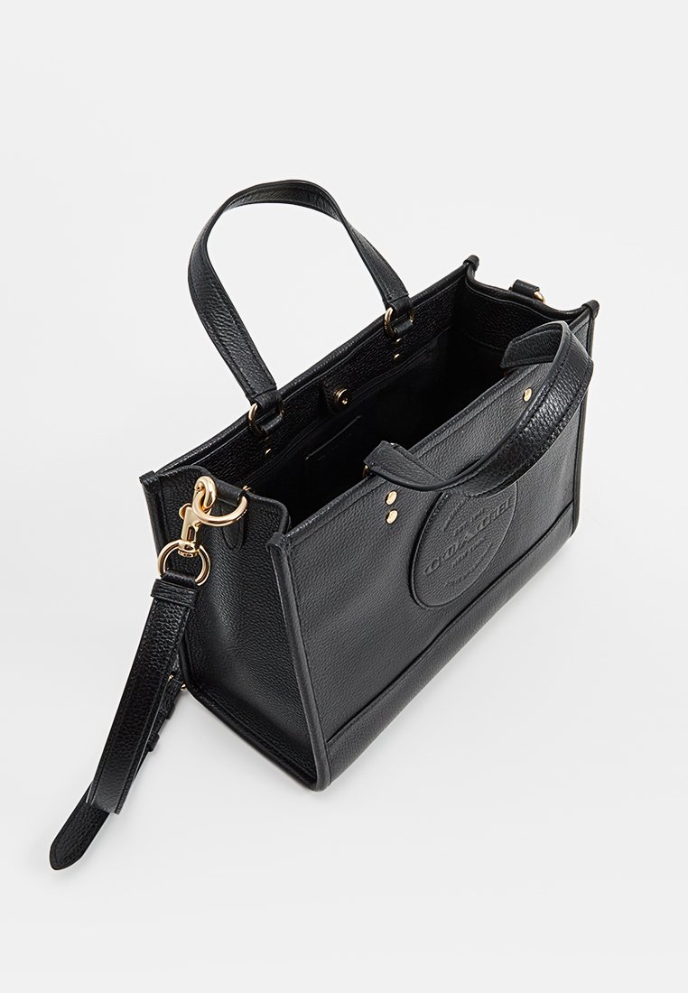 Bolso de mano de cuero negro con superficie texturizada, asas superiores y correa desmontable. Presenta herrajes dorados y un parche de logo circular en la parte frontal.