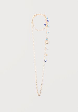 Collier en chaîne dorée avec un œil bleu maléfique, une perle et des perles en or espacées sur un côté, sur un fond uni.