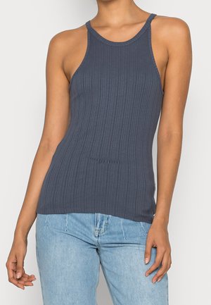 Kobieta w bezrękawniku o prążkowanej fakturze w ciemnoszarym kolorze i jasnoniebieskich dżinsach, stojąca na jasnym tle.