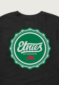 Camisa negra con un gran logotipo circular verde que presenta la palabra "Etnies", el texto "SKATEBOARDING" debajo y una pequeña "m" estilizada roja en la parte inferior.